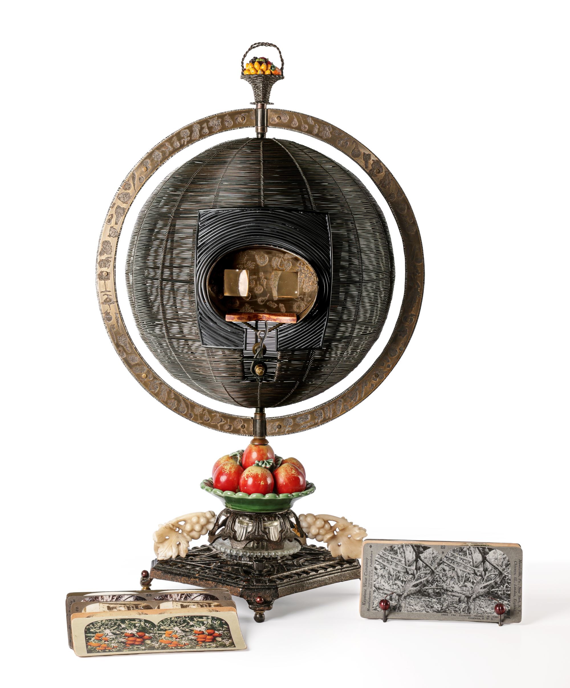 A globe-like sculpture with fruits and vegetables made out of steel, bronze, brass, marble, ceramic, and stereocards.